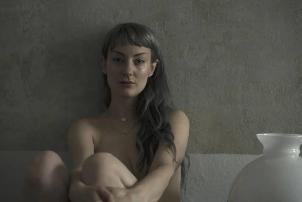 Vex Ashley assise contre un mur gris, les genoux repliés, près d’un vase blanc